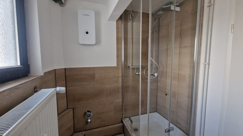 Modernes Badezimmer mit Glasdusche, Heizkörper und Bosch-Wasserboiler an der Wand. Helle Fliesen und Holzoptik.