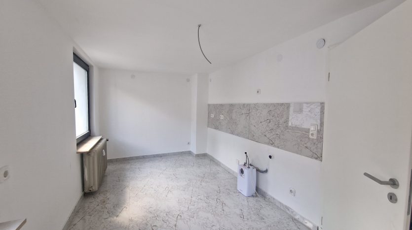 Helle, leere Wohnung mit marmoriertem Boden, Heizkörper an der Wand und großen Fenstern. Weiße Wände und Tür.