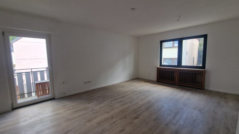 Helles Wohnzimmer mit Laminatboden, großer Fensterfront und Zugang zum Balkon. Heizkörper unter den Fenstern.