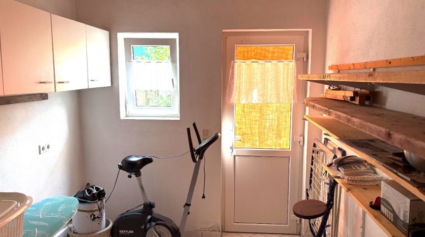 Küche mit Fenster, Tür, Holzregalen, einem Kettler Ergometer und einer Sitzgelegenheit. Helle Wände und Fliesenboden.