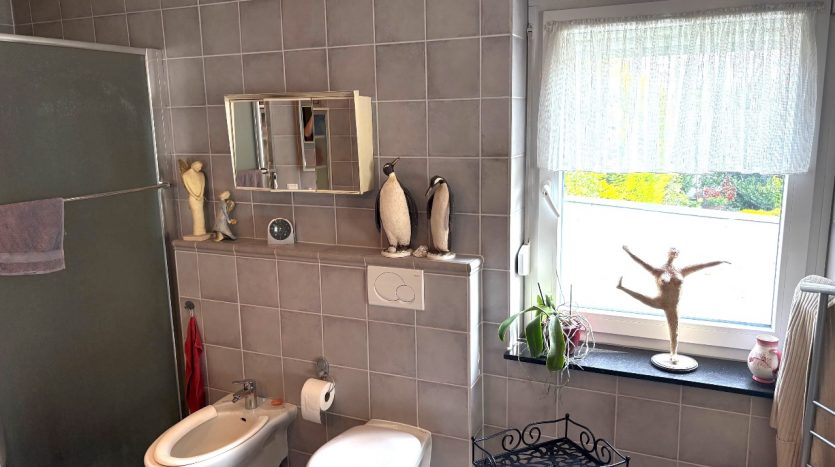 Modernes Badezimmer mit grauen Fliesen, Dusche, Bidet, Spiegel und dekorativen Figuren wie Pinguinen und einer Tänzerin.