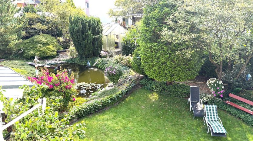 Blick auf einen Garten mit Teich, Blumen, zwei Liegen und einem Gewächshaus im Hintergrund.