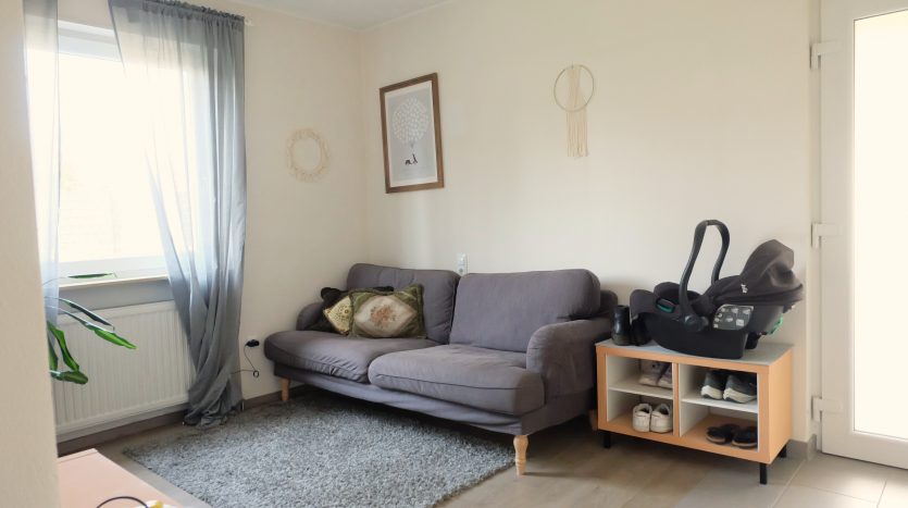 Heller Wohnraum mit grauem Sofa, dekorativen Kissen, Teppich und einem Kinderautositz auf einem Regal.