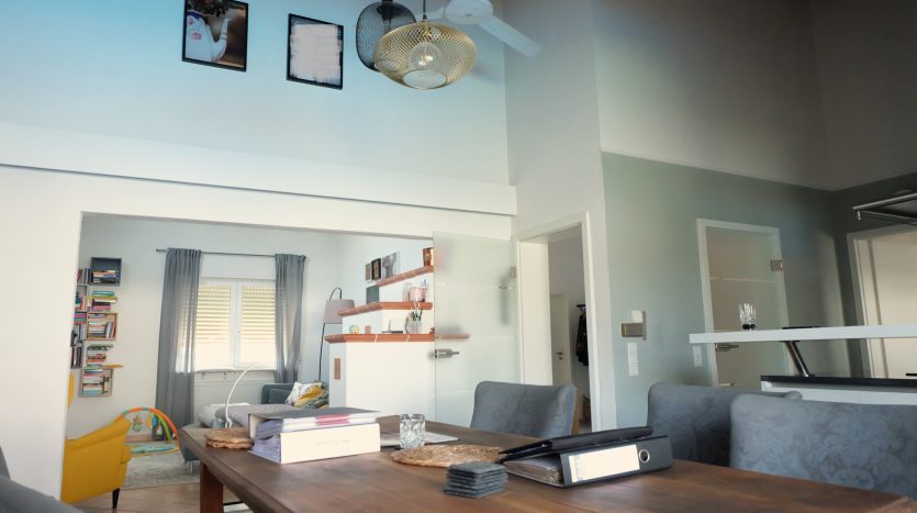 Helles Wohnzimmer mit Holztisch, grauen Stühlen, Bücherregal, gelbem Sessel und moderner Deckenlampe.