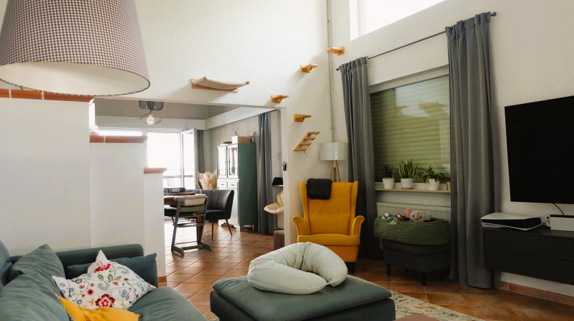Heller Wohnraum mit grünem Sofa, gelbem Sessel, Esstisch und großen Fenstern. Pflanzen und moderne Dekorationen sichtbar.