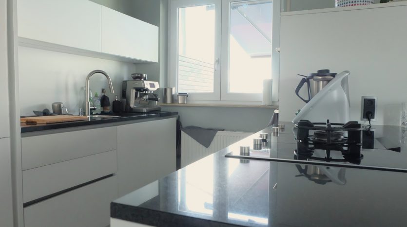 Moderne Küche mit weißen Schränken, schwarzer Arbeitsplatte, Kochfeld, Kaffeemaschine und Fenster.
