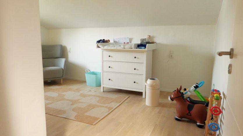 Helles Kinderzimmer mit einem grauen Sessel, einer weißen Kommode, einem Spielzeugpferd und buntem Spielzeug.