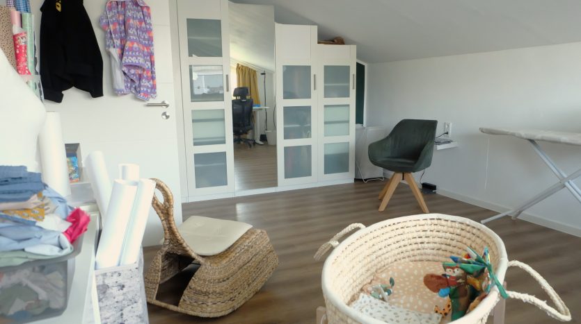 Heller Raum mit Holzfußboden, einem Rattanstuhl, einem Babybett und einem weißen Schrank mit Glastüren.