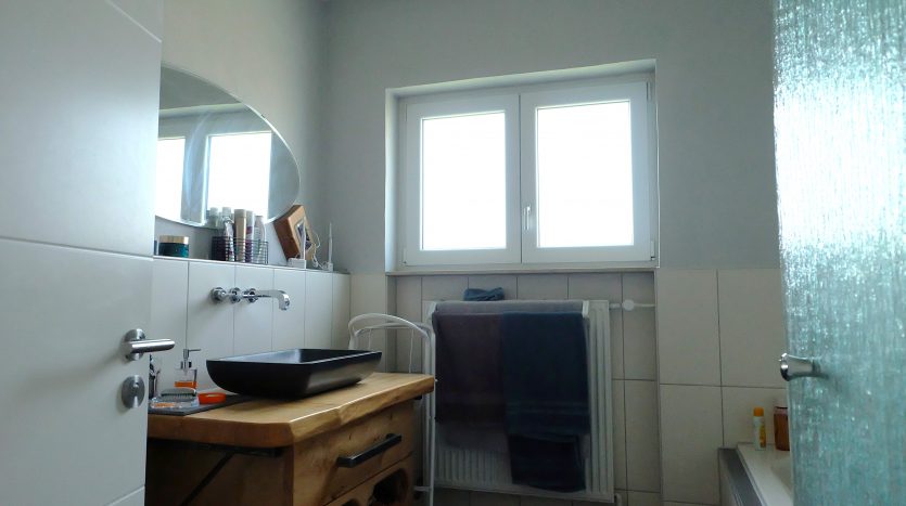 Helles Badezimmer mit einem Holzwaschtisch, schwarzem Waschbecken, Handtüchern und zwei Fenstern.