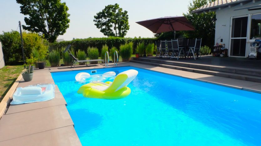 Pool mit klarblauem Wasser, bunten Luftmatratzen, Sonnenschirm und Gartenmöbeln im Hintergrund.