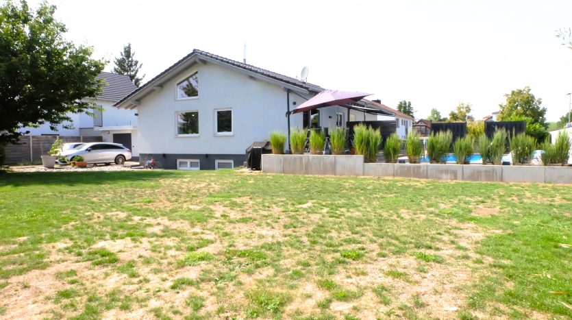 Einfamilienhaus mit Garten, Terrasse und Pool. Graue Wand, grüne Pflanzen und ein Auto im Vordergrund.