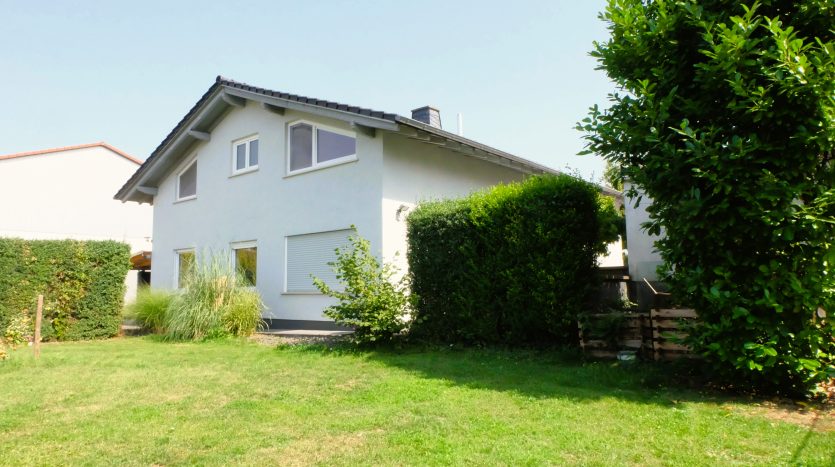 Zweifamilienhaus mit modernem, weißem Anstrich, Fenster mit Rollläden, umgeben von grünem Garten und Hecken.