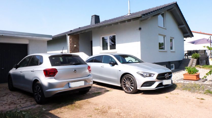 Zwei silberne Autos, ein VW und ein Mercedes, parken vor einem modernen, hellen Wohnhaus mit Garten.