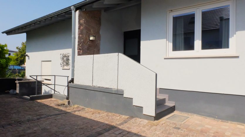 Eingangsbereich eines Hauses mit Treppe, grauer Fassade und Steinverkleidung, umgeben von gepflastertem Boden.