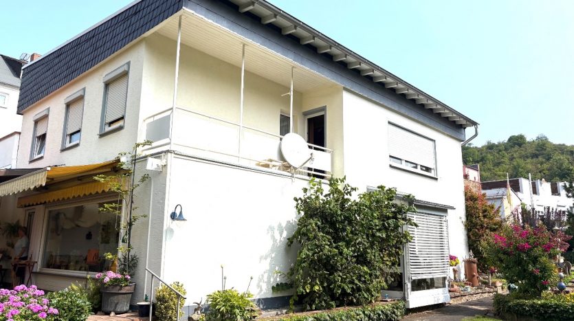 Zweistöckiges Wohnhaus mit Balkon, Satteldach und Satellitenschüssel, umgeben von bunten Blumen und Pflanzen.
