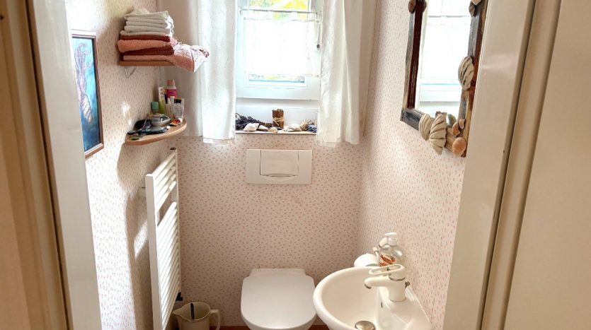Kleines Badezimmer mit Toilette, Waschbecken, Handtüchern, Wandspiegel und Fenster. Helle, freundliche Atmosphäre.