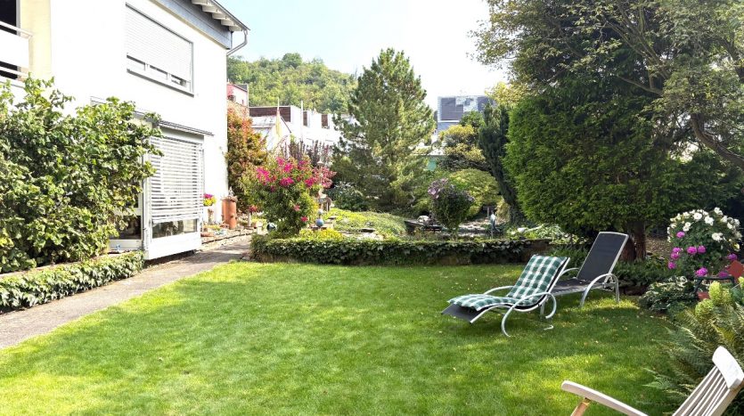 Blick auf einen gepflegten Garten mit grünem Rasen, Liegen und bunten Blumenbeeten neben einem modernen Haus.