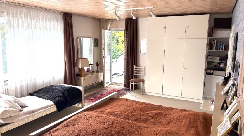 Helles Schlafzimmer mit Doppelbett, Tagesdecke in Braun, Schreibtisch, Spiegel und Balkonzugang.