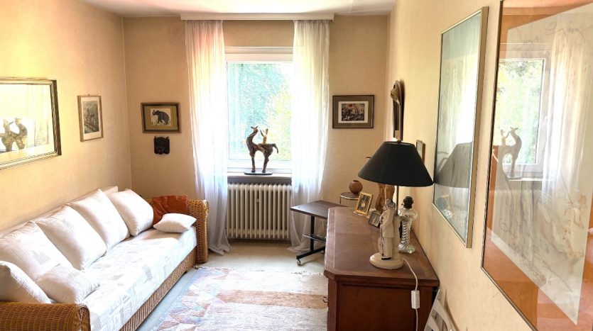 Wohnzimmer mit Sofa, Tischlampe und Holzmöbel. Kunstwerke an den Wänden, Fenster mit Vorhängen.