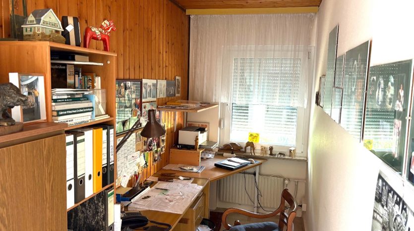 Büro mit Schreibtisch, Stühlen, Regalen und Bildern an der Wand. Holzvertäfelung und Fenster mit Vorhängen sichtbar.
