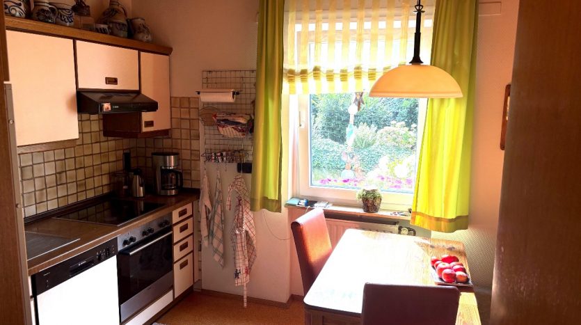 Kleine Küche mit Holzdecke, grünen Vorhängen, einem Tisch mit Stühlen und Küchengeräten wie Herd und Kühlschrank.