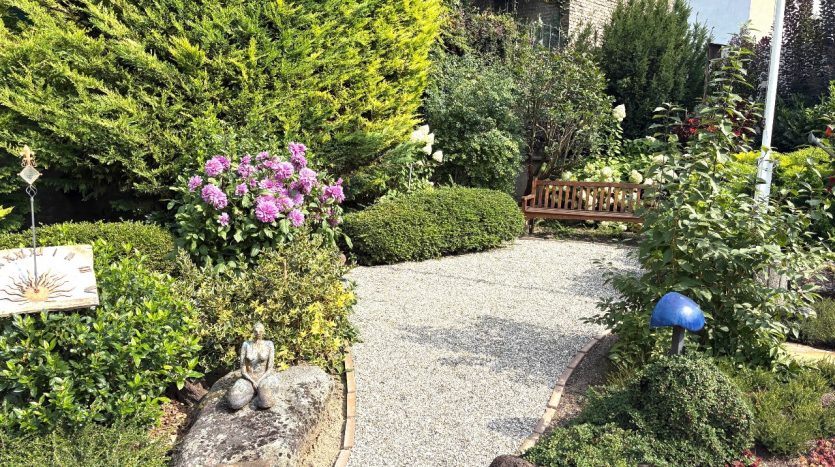 Blühende Hortensien, grüne Sträucher und ein gemütlicher Holzbank im gepflegten Garten mit Kiesweg.