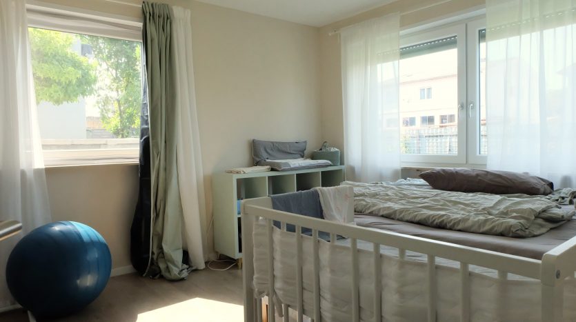 Helles Kinderzimmer mit einem Bett, einem Babybett, einem Wickeltisch und einem Gymnastikball. Fenster mit weißen Vorhängen.