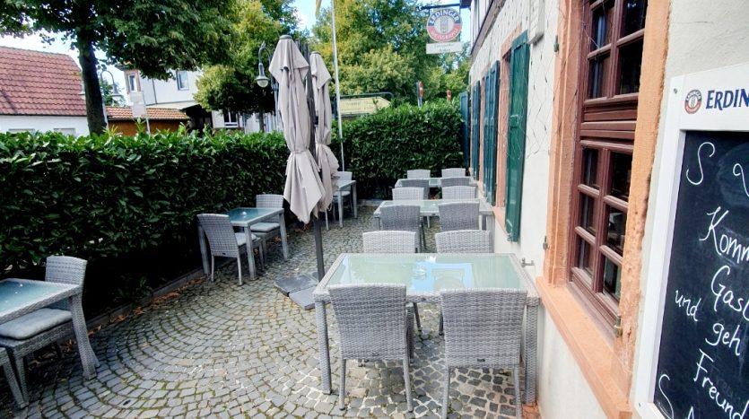 Gemütlicher Außenbereich mit grauen Sitzmöbeln, gepflastertem Boden und grünen Hecken. Sonnenschirme und Pflanzen dekorieren den Raum.