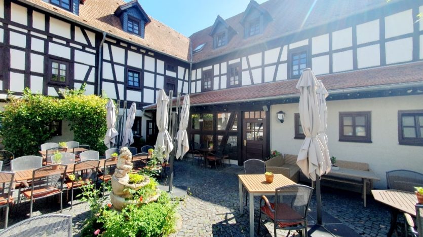 Gemütlicher Innenhof mit Steinpflaster, Tischen, Stühlen und Sonnenschirmen, umgeben von Fachwerkhäusern.