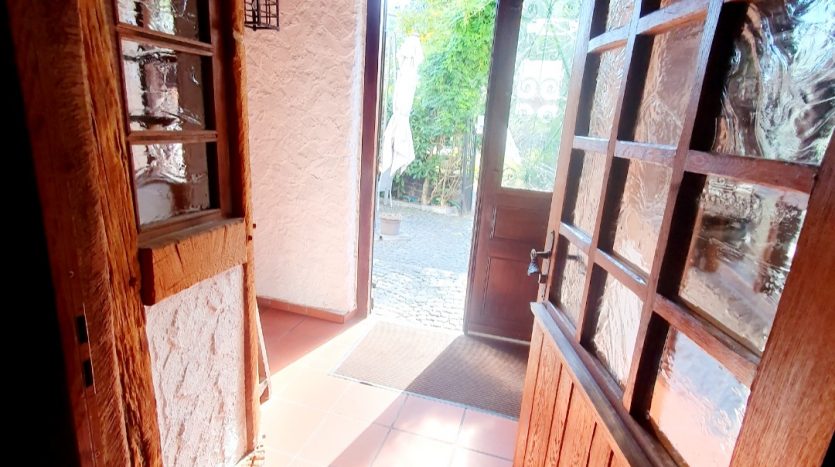 Blick durch eine offene Holztür in einen hellen Eingangsbereich mit Fliesenboden und Sicht auf den Außenbereich.