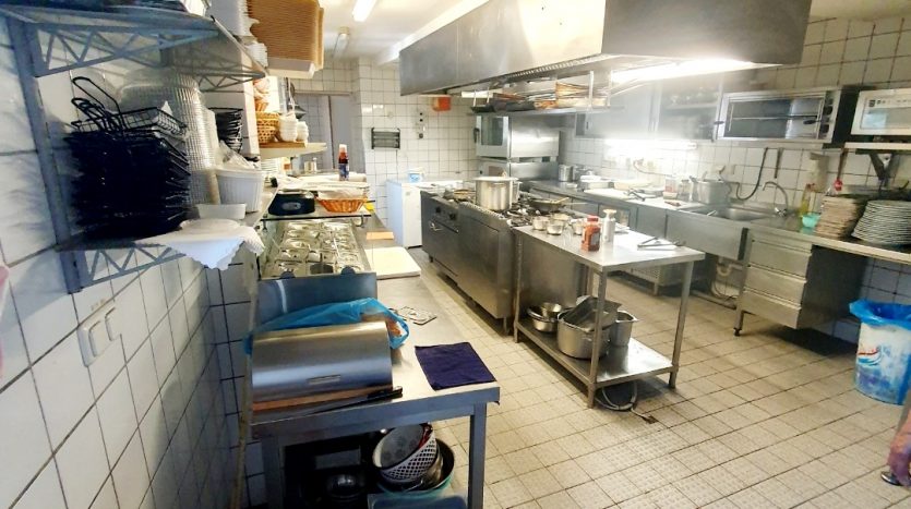 Gastronomieküche mit Edelstahlgeräten, Arbeitsflächen, Geschirr und Küchenutensilien, gefliestem Boden.