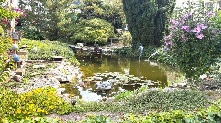 Idyllischer Garten mit Teich, Seerosen, Koi-Fischen, Pflanzen, Skulpturen und blühenden Sträuchern.