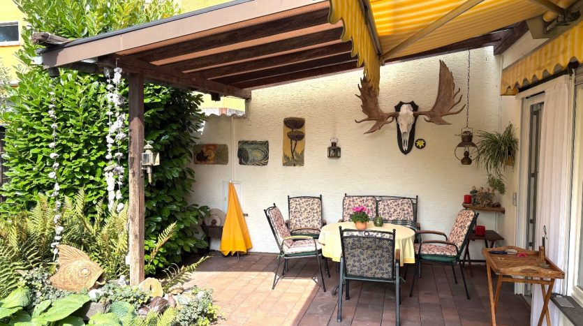 Gemütliche Terrasse mit Holzpergola, Tisch und Stühlen, umgeben von Pflanzen und Dekorationen, darunter ein Geweih.