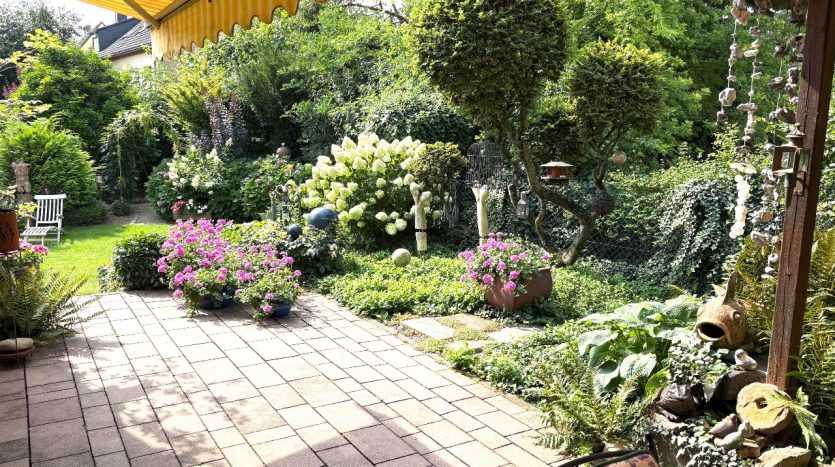 Blühender Garten mit bunten Blumen, skulpturalen Elementen und gepflegtem Rasen unter gelber Markise.